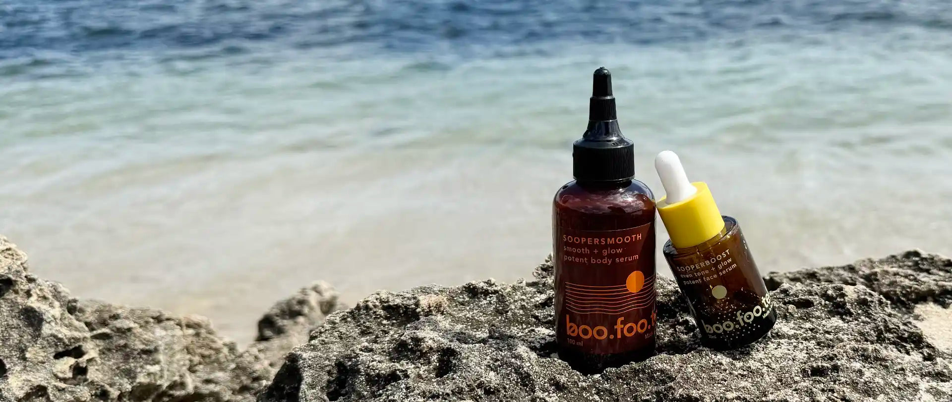 Two corrective serums for dark spots and bumps — Sooperboost face serum and Soopersmooth body serum by Boofootel, placed on rocks by the beach